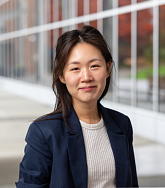 A smiling woman with dark hair wearing a white shirt and blue blazer poses in front of a wall of windows
