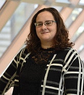 Person wearing a black lace top and a black-and-white patterned cardigan, standing indoors near large angled windows.