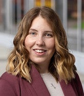 Person wearing a burgundy blazer over a light top, standing outdoors near a modern glass building.