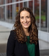 Smiling woman with long, curly hair wearing a black blazer over a green top, standing outdoors near a modern glass building.
