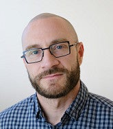 Man wearing a blue plaid button-up shirt against a plain, light-colored background.