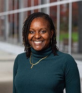 A woman wearing a dark green turtleneck and gold necklace, standing outdoors near a modern glass building.