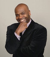 A smiling man wearing a black suit and striped shirt poses with his hand on his chin