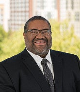 A man wearing glasses, a dark suit, and a tie smiles outdoors with green trees and tall buildings in the blurred background.