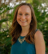 Kelsey Dickson, smiling, with long auburn hair, wearing a teal sleeveless top and statement necklace, outdoors with green foliage behind her.