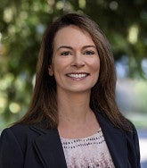 Karen Chaput smiling, with long brown hair, wearing a black blazer over a patterned blouse, outdoors in front of blurred greenery.