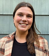 Madeleine Allen smiling, with long brown hair and a plaid flannel jacket over a black top, in front of gray wood siding.