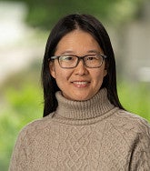 Yu Zhang smiling, with long black hair and glasses, wearing a tan cable-knit turtleneck sweater against a blurred green outdoor background.