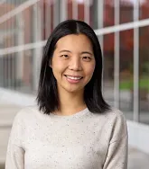 Headshot of a woman with straight black hair smiling outdoors in front of a modern campus building